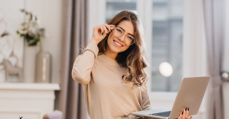 Cute girl in beige shirt touching glasses and holding laptop with smile. Indoor photo of beautiful female student preparing for lesson with computer at home..