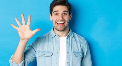 Close-up of handsome man smiling, showing fingers number five, standing over blue background