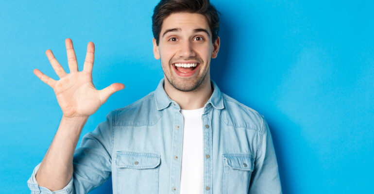 Close-up of handsome man smiling, showing fingers number five, standing over blue background