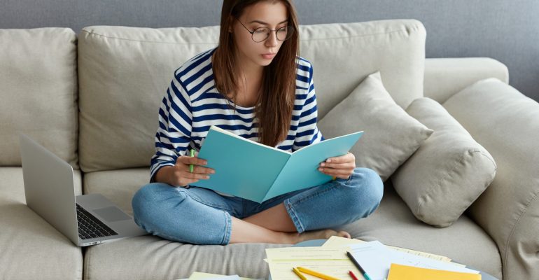 Young freelancer does ditance job at home, studies literature, learns foreign language in internet, writes down unknown words, sits crossed legs on couch, spends time at home, connected to wifi